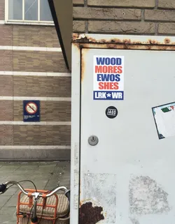 Street sticker The sticker is rectangular with a white background and features bold, sans-serif text arranged in columns. The top section has three lines: 'WOOD' in blue, 'MORES' in red, and 'EWOS' in blue. Below this, 'SHES' is in red. The bottom line reads 'LRK*WR' in blue with a red asterisk separating 'LRK' and 'WR'. The sticker appears to be placed on a light-colored, possibly metal surface that shows signs of rust and wear.