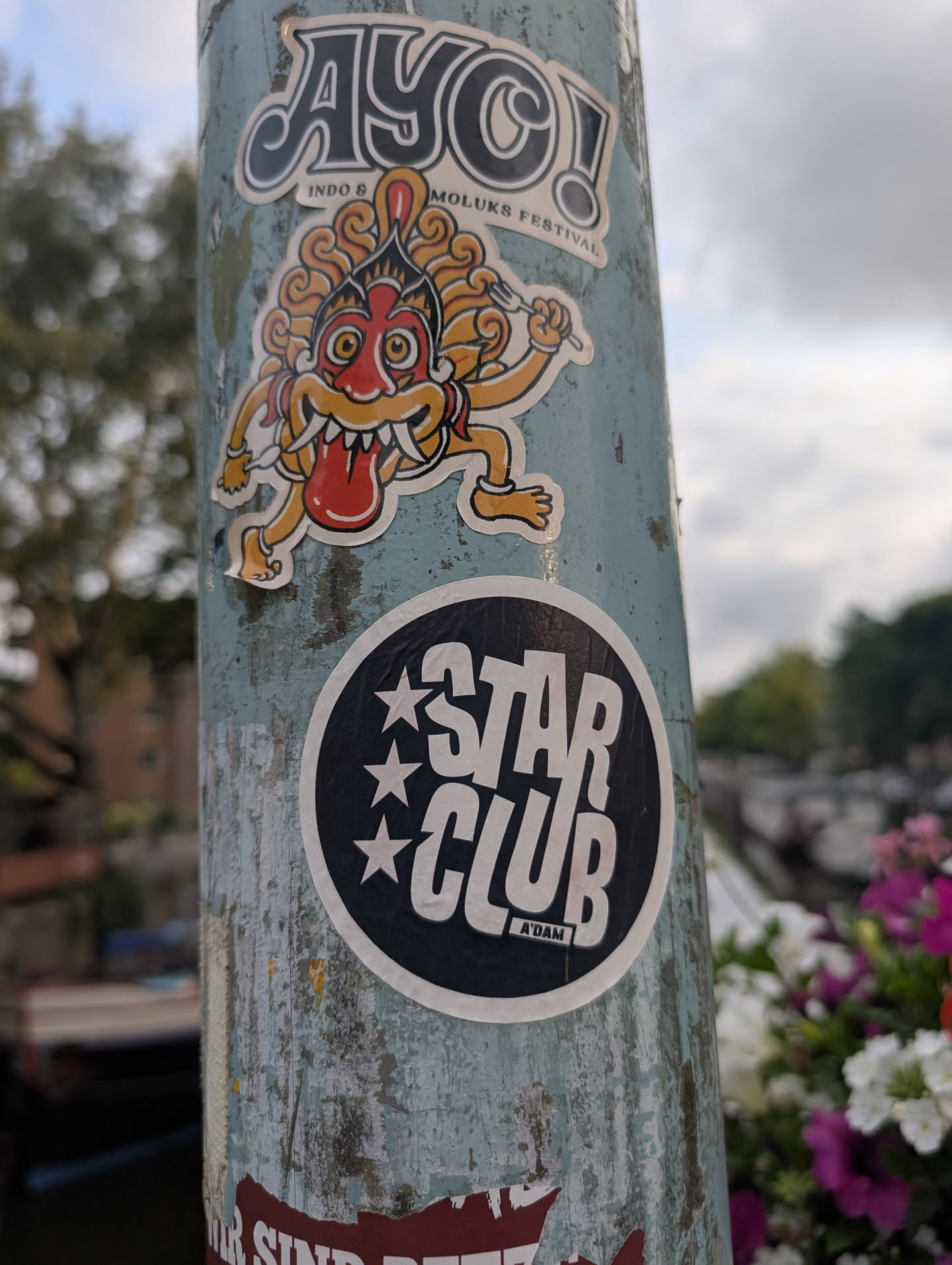 The image displays two stickers on a weathered, pale blue pole. The top sticker features a brightly colored, stylized creature resembling a Barong or a similar mythological entity from Indonesian culture. It has a red face with a gaping mouth, yellow eyes, and ornate orange and yellow headdress with red accents. The creature is depicted with sharp teeth and a long, red tongue sticking out. It is holding a fork in its right hand and has simplified limbs. Above this creature, in bold, stylized lettering with a black outline and a pale yellow fill, is the text 'AYO!'. Below 'AYO!', in smaller text, it reads 'INDO & MOLUKS FESTIVAL'. The second sticker, positioned below the creature, is circular with a thick black border and a white inner circle. Inside the white circle, the words 'STAR CLUB' are prominently displayed in bold, white, sans-serif lettering. Below 'STAR CLUB', in smaller white text, is 'A'DAM'. Three white stars are arranged vertically to the left of 'STAR CLUB'. Both stickers show signs of being affixed to the pole for some time, with some wear and tear visible around the edges.