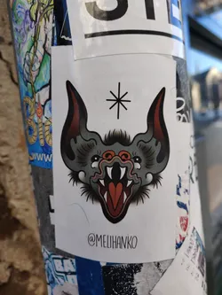 A sticker featuring a traditional tattoo-style illustration of a bat's head with its mouth open, showing sharp teeth. The bat is grey and red. A small asterisk is above its head.  The artist's Instagram handle is printed at the bottom of the sticker.