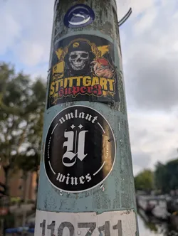 Street sticker The sticker at the center of the image is circular with a black background. It features a large, stylized Gothic letter 'U' in white. Around the letter, the text "amsterdam" is printed in an arc on the left side, and "wines" is printed in an arc on the bottom. On the right side, "EST. MMXXIV" is visible in smaller print. The sticker has a white border.