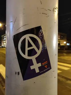 A sticker with a symbol combining the anarchy symbol with the female gender symbol. It also contains the text "wijebond" and a website address.
