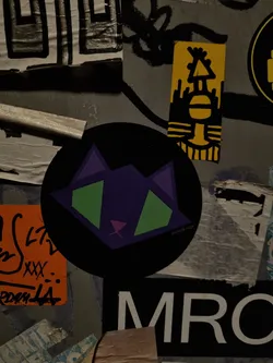 Street sticker A round black sticker with a geometric design of a cat in purple, green, and pink. The artist's Instagram handle is @vivid.lugg.