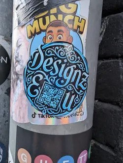 Street sticker A round black sticker featuring stylized cyan calligraphy spelling 'Designz & u'. The text is embellished with elegant swirls and scrollwork. A matching cyan QR code is integrated into the design on the lower left.