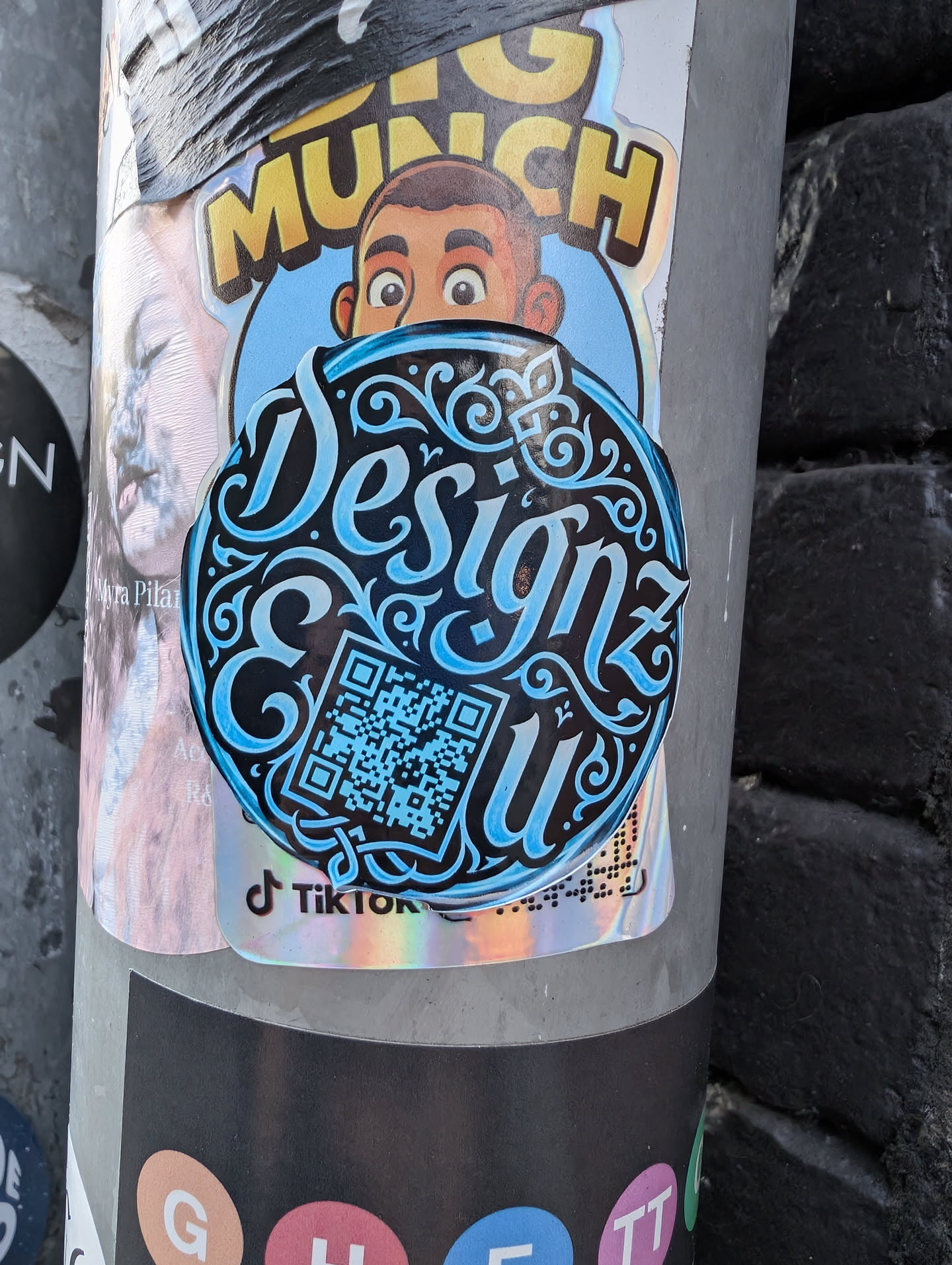 A round black sticker featuring stylized cyan calligraphy spelling 'Designz & u'. The text is embellished with elegant swirls and scrollwork. A matching cyan QR code is integrated into the design on the lower left.