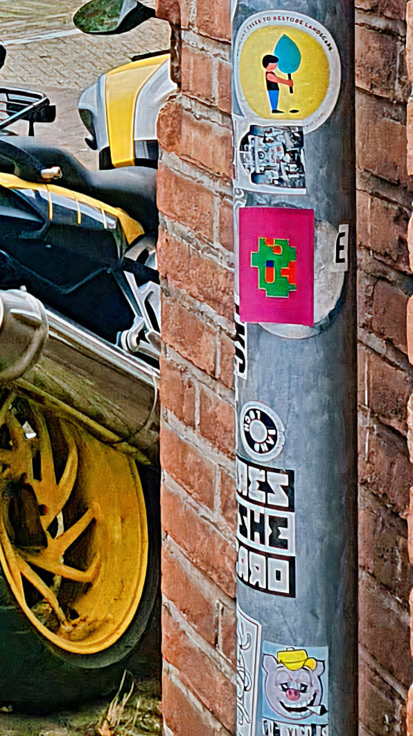 The central sticker is a bright pink square featuring a pixelated design composed of green and orange elements. It's affixed to a metal pole which also bears several other stickers of varying designs. The pole stands next to a brick wall on one side and a motorbike on the other.