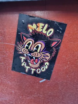 Street sticker A black sticker with a cartoon cat illustration. The text "MELO" is written above the cat, and "TATTOOS" is written below the cat. The cat's eyes are wide, and it has a large mouth with sharp teeth. The sticker has a slightly worn appearance.
