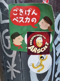 Street sticker A collection of stickers on a dark surface. The main sticker features a speech bubble in red with Japanese text on a green background. There are two simple cartoon characters, one speaking in Japanese.  A smaller sticker underneath displays a cartoon man with blond hair, the word "Arsch" prominently displayed, and text in German around the edges.