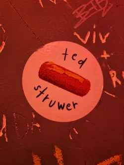 Street sticker A round sticker with a drawing of a fried food, and the text "ted strüwer" written on it. The sticker is attached to a dark red wall with other graffiti around it.