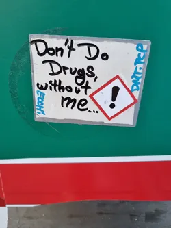 Street sticker A white square sticker with black handwritten text that says: Don't Do Drugs, without me... There is also a small red and white warning sign with an exclamation mark. The sticker is placed on a green surface with a red stripe below.