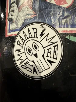 Street sticker A circular, black and white sticker featuring a stylized, cartoonish skull or monster head at its center. The head has large, wide-set black eyes and a small, inverted triangular nose. Lightning bolt shapes emanate from the sides of the head. Arched over the top of the head are the letters 'RAAAR'. On the left side, vertically oriented, are the letters 'MESS', and on the right side, horizontally, are the letters 'RE'. The entire design is rendered in a thick, hand-drawn, somewhat crude style with a black outline and details on a white background.