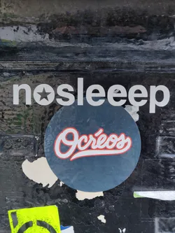 Street sticker Round sticker with the text "Ocréos" in a stylized red and white font on a dark blue background.  The sticker is placed on a dark surface with other stickers and markings.  Above the sticker is a banner with the text "nosleep" in a sans-serif font.