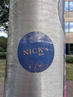Street sticker A round blue sticker on a metal post. It displays 'NICK'S BAR' in orange text, flanked by orange line drawings of cherubs—one resting at the top, and one holding a glass and smoking at the bottom.
