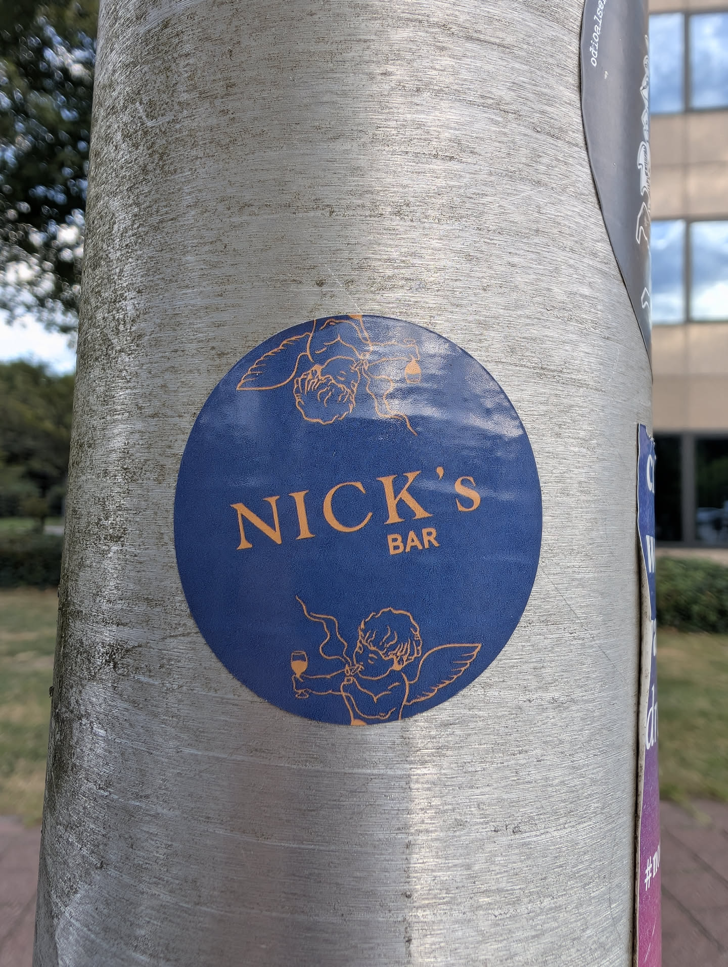 A round blue sticker on a metal post. It displays 'NICK'S BAR' in orange text, flanked by orange line drawings of cherubs—one resting at the top, and one holding a glass and smoking at the bottom.