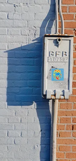 A small pixelated art sticker adorns an electrical box mounted on a brick wall. The sticker features a design of various colors including red, yellow and blue, reminiscent of early computer graphics. The metal box shows signs of weathering, contrasting with the brightly colored sticker.