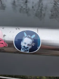 Round sticker featuring a photo of a person with light skin and short brown hair.  They are wearing what appears to be a donkey mask or filter over their face and light blue donkey ears are digitally added to the top of their head. The background of the sticker is dark blue. The sticker is affixed to a metal railing outdoors. 