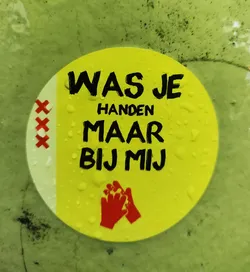 Street sticker A yellow sticker with the text "Was je handen maar bij mij" (Wash your hands, but with me) in black. It also features a pictogram of two hands washing each other and three red crosses on the left side. The sticker appears to be affixed to a green surface with water droplets visible on it.
