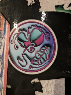 A circular sticker featuring a highly stylized, abstract face or creature design. The sticker has a white outer border, followed by a bright magenta inner border. The background of the central design transitions from a light sky blue on the left to a darker purple-blue on the right. The main elements consist of two large, oval-shaped 'eyes' or eye-like features filled with a bright teal/aqua color, outlined in magenta. Below these, a complex, web-like or skeletal structure, also in teal/aqua with magenta outlines, suggests a mouth or lower jaw. Numerous magenta lines and swirls emanate from and surround these central features, creating a dynamic, organic, and somewhat gooey or liquid appearance. The sticker has a glossy finish, reflecting light in several areas, and is applied to a dark, textured surface with other torn paper fragments visible.