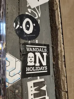 A black and white sticker with the text "VANDALS ON HOLIDAYS" and a smiley face wearing a balaclava. The sticker is affixed to a wall.