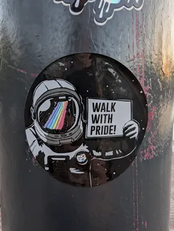 Street sticker Circular black sticker depicting an astronaut with a rainbow reflected in their visor, holding a sign that reads 'WALK WITH PRIDE!'.