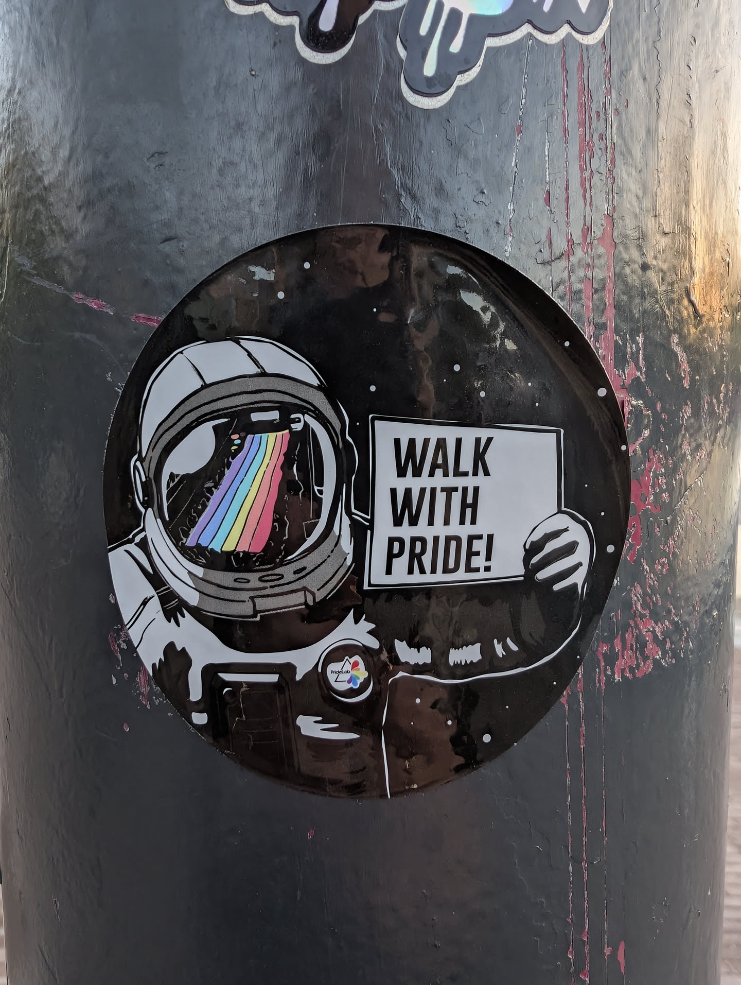 Circular black sticker depicting an astronaut with a rainbow reflected in their visor, holding a sign that reads 'WALK WITH PRIDE!'.