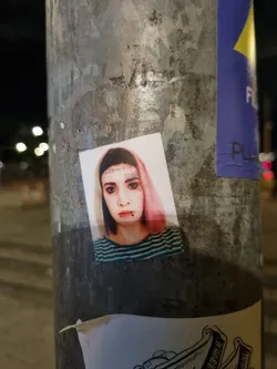 A sticker featuring a close-up portrait of a young woman with pink hair, facial piercings, and a striped shirt. The sticker is affixed to a weathered metal pole outdoors.