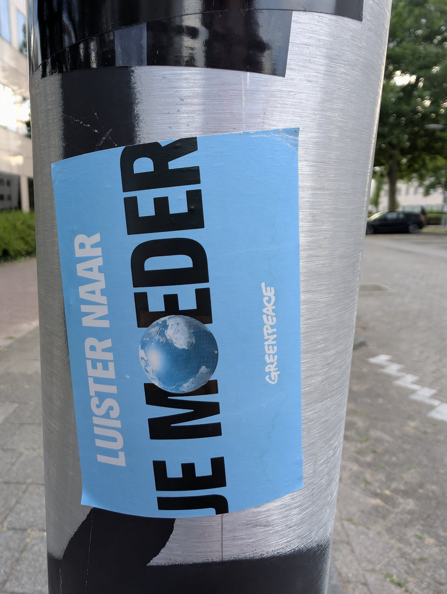 A blue rectangular sticker pasted vertically on a metal pole. It features the Dutch text 'LUISTER NAAR JE MOEDER' in white and black, with a globe graphic replacing the letter 'O' in 'MOEDER'. The Greenpeace logo is printed on the side.