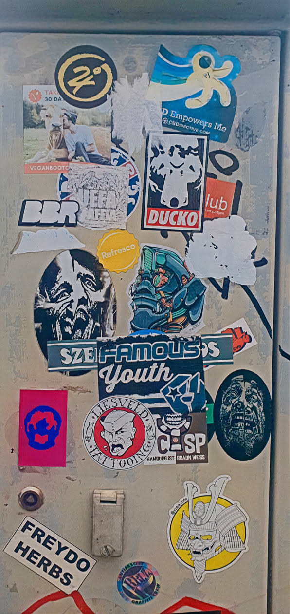 This image showcases a cluster of stickers with varied themes and styles on a weathered surface. The stickers range from artistic designs to logos and textual elements, creating a visually rich and layered composition. Some stickers are peeling or damaged, adding to the aged effect.