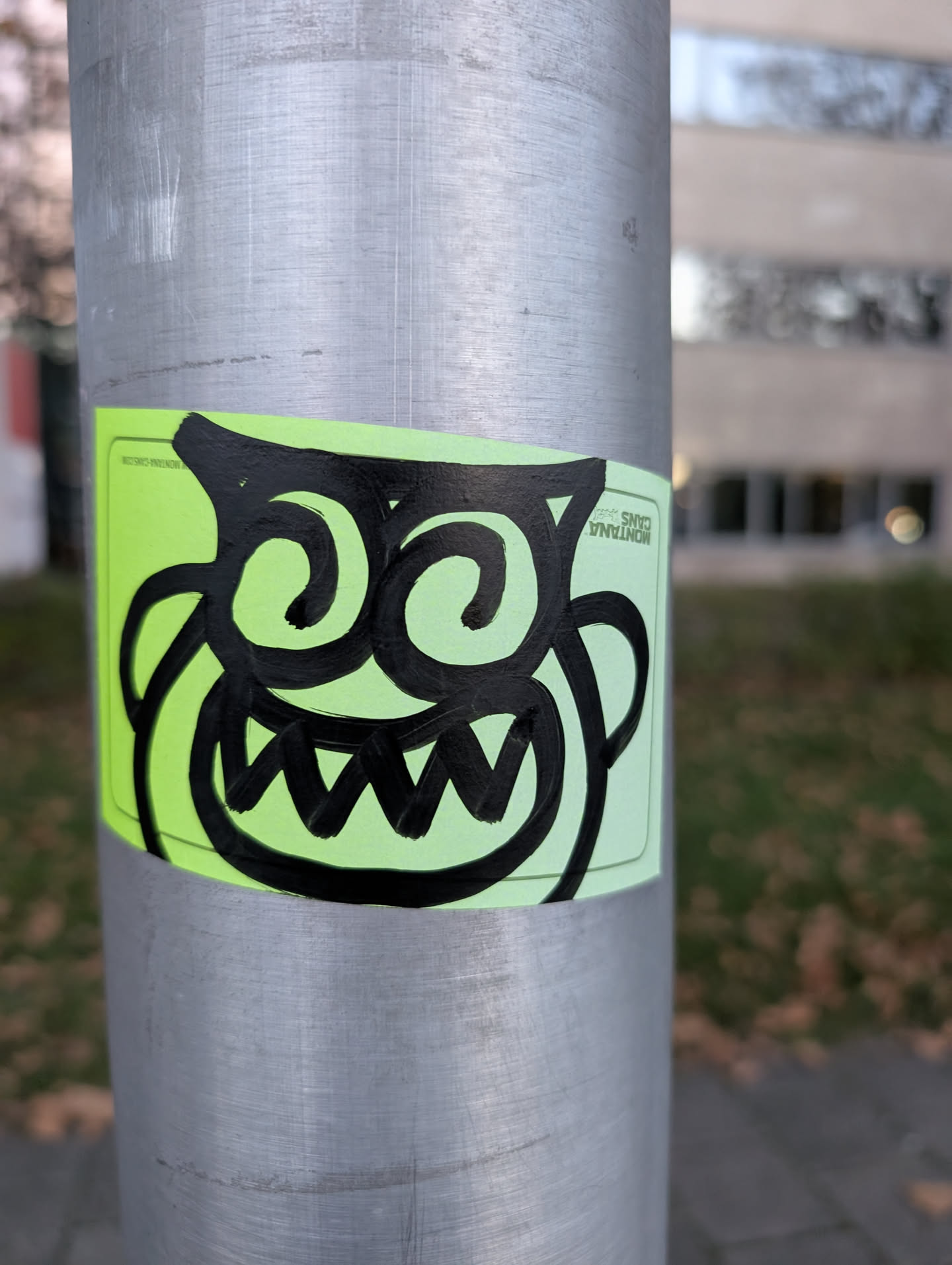 The sticker is rectangular with rounded corners and has a bright neon green background. Centered on the sticker is a stylized, cartoonish monster face drawn in thick, bold black lines. The monster has large, swirling eyes that resemble question marks or spirals, and a wide, jagged mouth filled with sharp, triangular teeth. Its face is framed by two large, curved lines that could be interpreted as ears or horns. The overall impression is energetic and slightly mischievous.