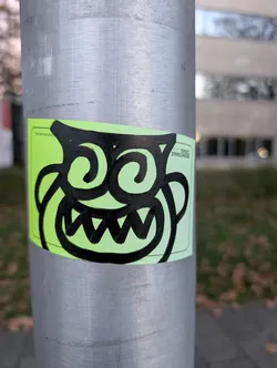 Street sticker The sticker is rectangular with rounded corners and has a bright neon green background. Centered on the sticker is a stylized, cartoonish monster face drawn in thick, bold black lines. The monster has large, swirling eyes that resemble question marks or spirals, and a wide, jagged mouth filled with sharp, triangular teeth. Its face is framed by two large, curved lines that could be interpreted as ears or horns. The overall impression is energetic and slightly mischievous.