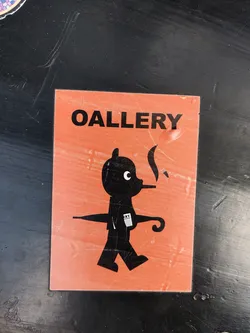 Street sticker This sticker features a unique graphic design. A stylized figure with an umbrella and a cigarette is the focal point.  The color scheme is simple: black illustrations on an orange background. The word "OALLERY" at the top is a playful misspelling of "GALLERY", adding to its quirky aesthetic.  The sticker appears to be slightly worn, hinting at its placement in an outdoor environment. 
