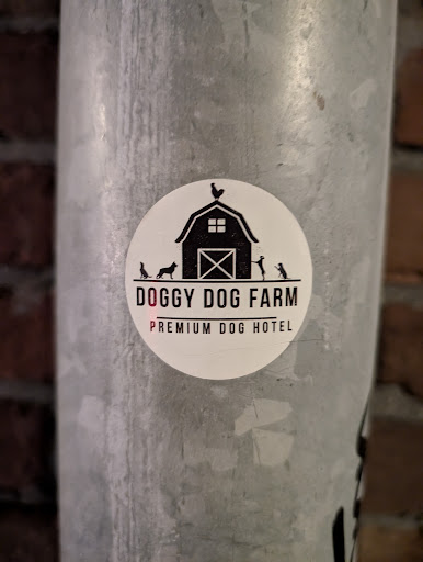 A round sticker on a metal pole advertises "Doggy Dog Farm", a premium dog hotel. The design features a black barn silhouette with a rooster on top, flanked by dog and human figures.