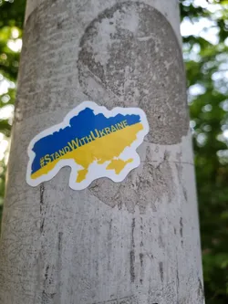 Street sticker A sticker expressing solidarity with Ukraine, featuring a map of the country in its national colors (blue and yellow).  The sticker is affixed to a gray pole outdoors.