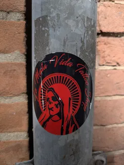 Street sticker A round sticker with a black background and a red image of a skull-like figure resembling the Virgin Mary with a halo. The text 'La Mala Vida Tattoo Shop' curves around the top edge of the sticker.