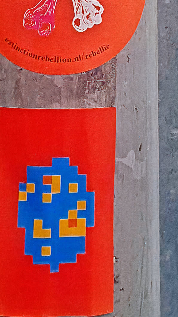 A sticker featuring a blue and yellow pixelated alien-like figure on a square red background. The pixel art is reminiscent of old arcade games. Above it, partially visible, is a round sticker with an illustration and the text: extinctionrebellion.nl/rebellie