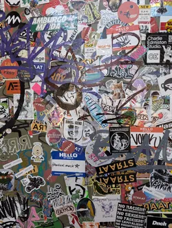 A close-up shot of a wall covered in a multitude of stickers. The central sticker displays the text "Radical mask".  Many other stickers have illegible text, while some show logos and imagery. The overall aesthetic is one of urban grit and artistic expression. 