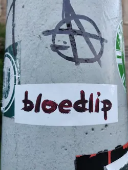 Street sticker A white rectangular sticker with the word \"bloedip\" written in dark red paint. The lettering style is slightly irregular, giving it a hand-painted look.  The sticker is affixed to a gray pole, which also has other graffiti and stickers on it.