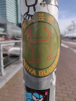 A round green sticker with an orange outline featuring a stylized image of a female warrior's (onna bugeisha) helmet with bull horns. The text 'ONNA BUGEISHA' is written in orange around the central image.  Japanese characters are also visible on the sticker, adding to its unique design.