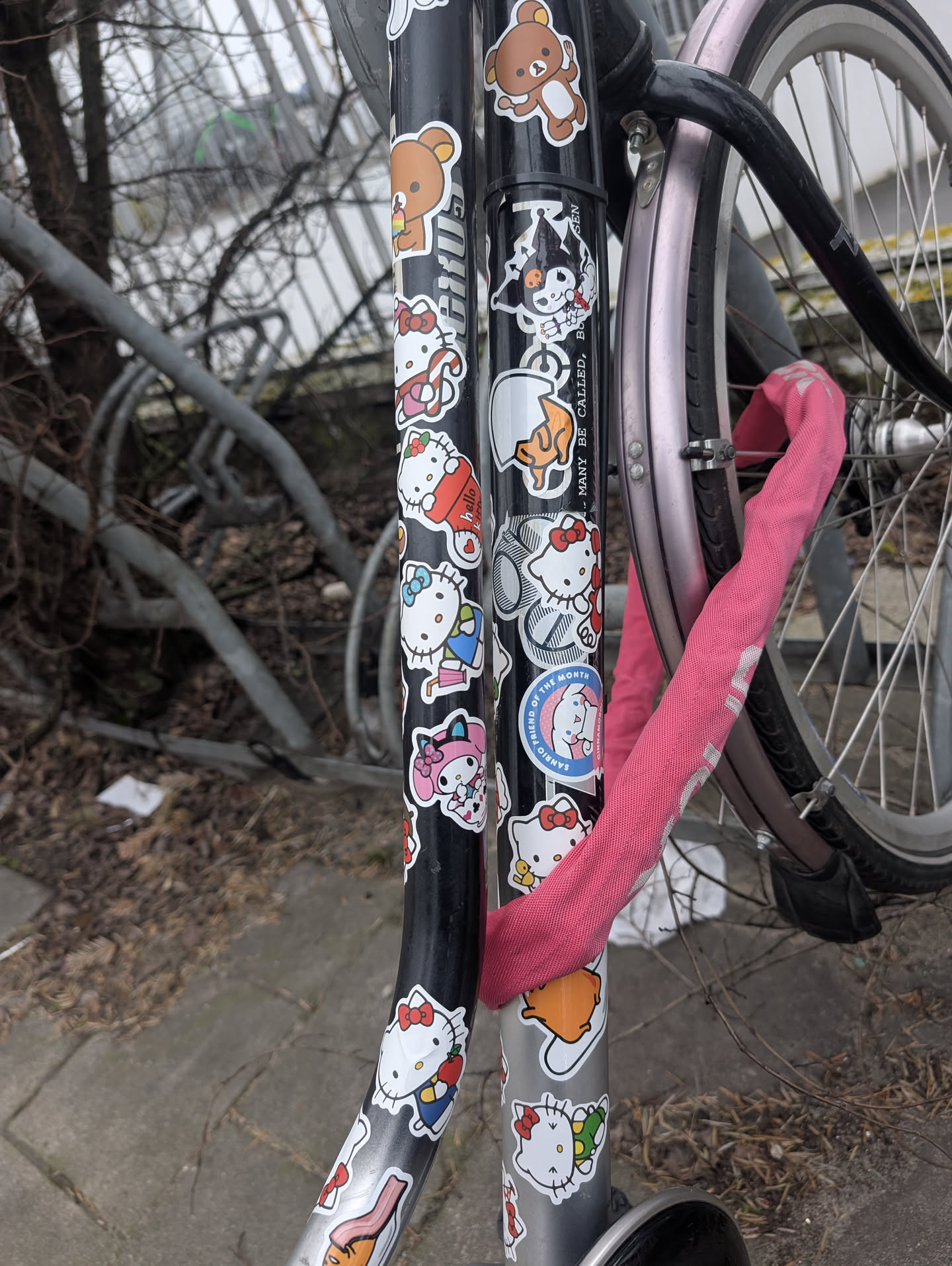 Street sticker The central image features a dense collection of colorful cartoon character stickers applied to the black and silver frame of a bicycle. The stickers are predominantly white with vibrant illustrations, showcasing a variety of popular Sanrio characters such as Hello Kitty (depicted in multiple poses and outfits, holding items like an apple, a red cup, or a present), My Melody, Kuromi, Cinnamoroll, and Gudetama (the lazy egg). A Rilakkuma bear sticker is also visible at the top. The stickers vary in size and are arranged in a somewhat overlapping, collage-like fashion. The bicycle frame is partially obscured by a bright pink chain lock. The overall appearance is cheerful and whimsical.