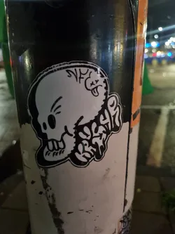 Street sticker A black and white sticker of a skull with some graffiti-style text and a smaller character on its head. The style is edgy and slightly cartoonish.