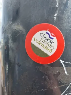 Street sticker A round sticker with a red border and a white center.  The text 'Cheese Factory Volendam' is written on it, along with a small image of the Dutch flag.