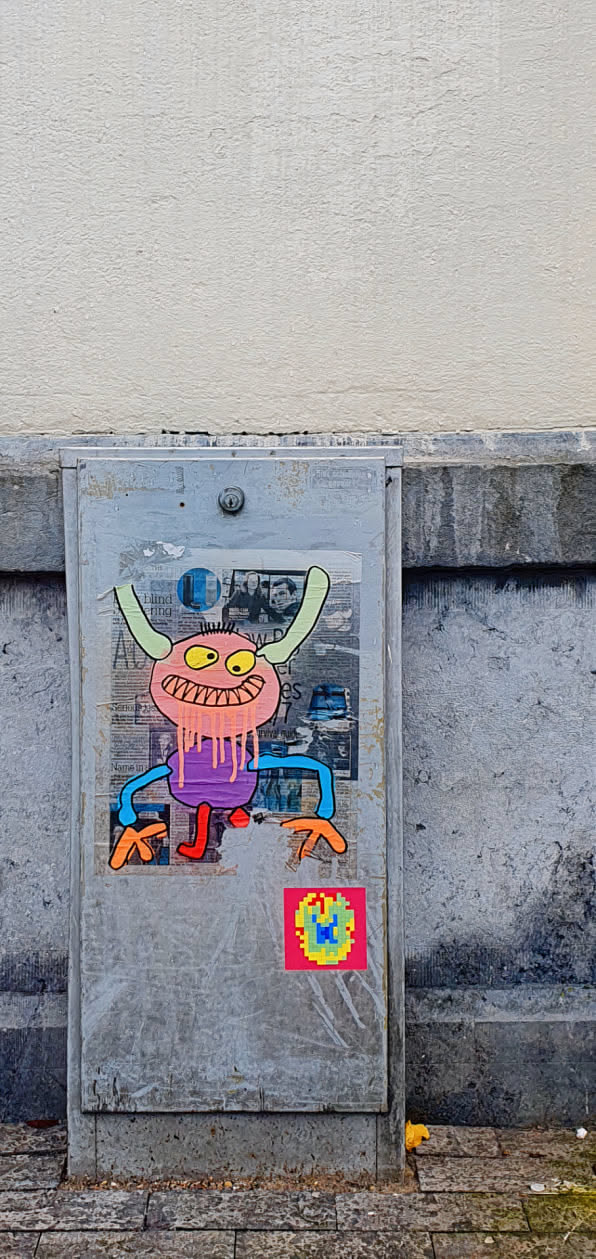 The sticker features a cartoonish monster with a pink head, yellow eyes, and a wide grin. It has light green horns, a purple body with dripping pink goo, and blue arms with orange hands. Its legs are red. There is a colorful, pixelated sticker beneath it.