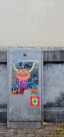 Street sticker The sticker features a cartoonish monster with a pink head, yellow eyes, and a wide grin. It has light green horns, a purple body with dripping pink goo, and blue arms with orange hands. Its legs are red. There is a colorful, pixelated sticker beneath it.