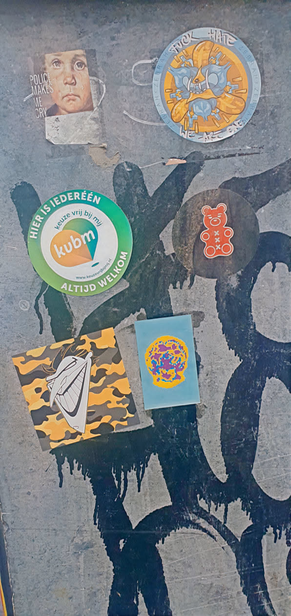 A collection of stickers on a weathered wall. One sticker features a crying child with the text "POLICE MAKES ME CRY". Another round sticker says "FUCK HATE". A green round sticker promotes 'keuze vrij bij mij' with a website address. Other stickers include a red gummy bear, a yellow camo pattern with a white grinning face, and a blue sticker with a pixelated image.