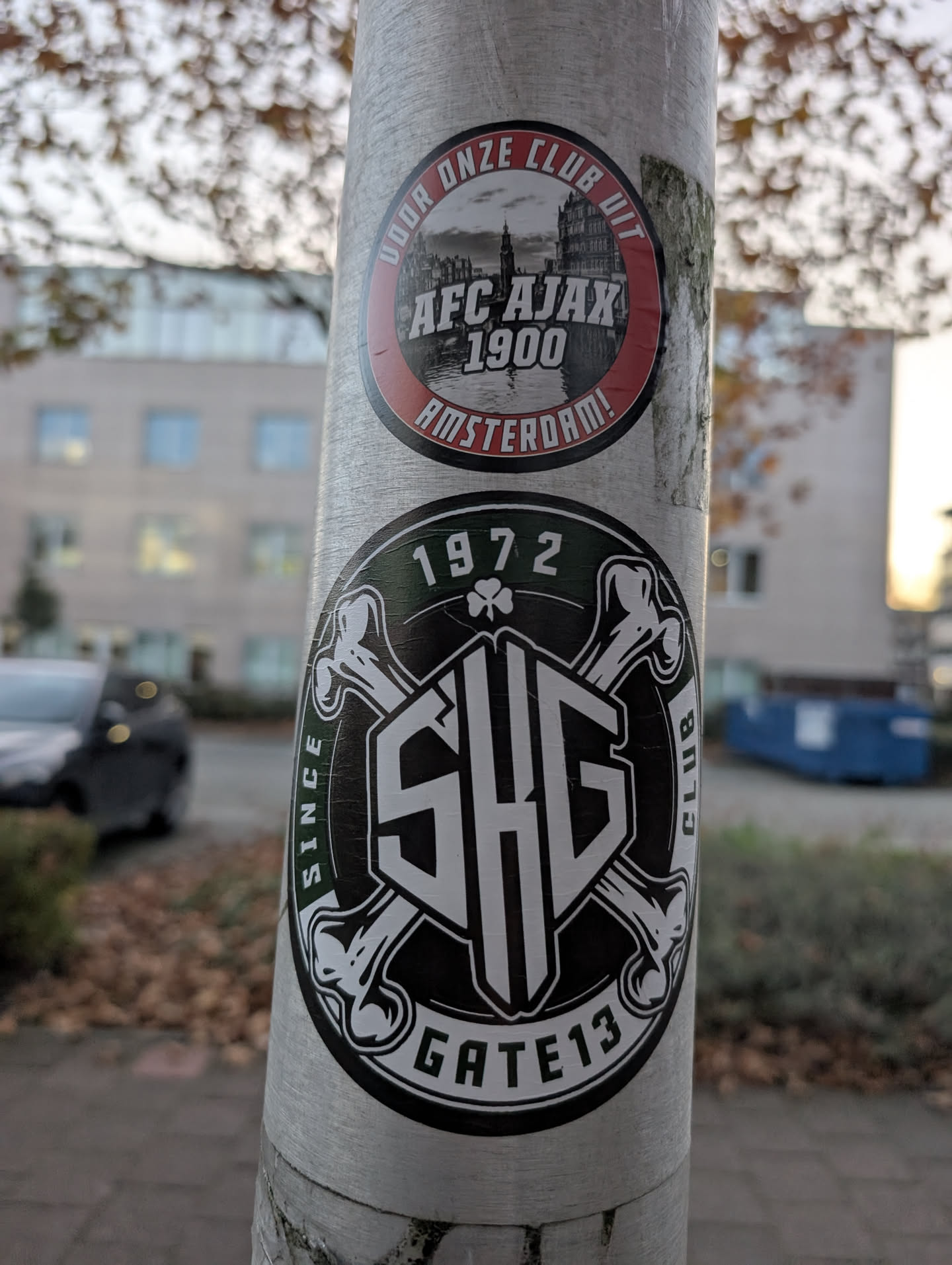 Street sticker The image features two circular stickers on a metal pole. The top sticker is red and black, with the text 'DOOR ONZE CLUB UIT' around the top edge, 'AFC AJAX 1900' in the center, and 'AMSTERDAM!' along the bottom edge. It also contains a black and white image of a cityscape with a prominent tower. The bottom sticker is black and green, with '1972' at the top, a shamrock symbol, and the letters 'SHG' in a stylized font in the center, surrounded by crossbones. The text 'SINCE' is on the left, 'CLUB' on the right, and 'GATE 13' along the bottom edge.
