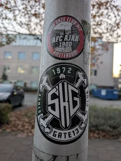 Street sticker The image features two circular stickers on a metal pole. The top sticker is red and black, with the text 'DOOR ONZE CLUB UIT' around the top edge, 'AFC AJAX 1900' in the center, and 'AMSTERDAM!' along the bottom edge. It also contains a black and white image of a cityscape with a prominent tower. The bottom sticker is black and green, with '1972' at the top, a shamrock symbol, and the letters 'SHG' in a stylized font in the center, surrounded by crossbones. The text 'SINCE' is on the left, 'CLUB' on the right, and 'GATE 13' along the bottom edge.
