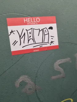 Street sticker A red name tag sticker with black graffiti-style lettering. The text on the sticker says "HELLO MY NAME IS:" followed by the stylized name KIOZA and a small tag HENXS at the bottom. 