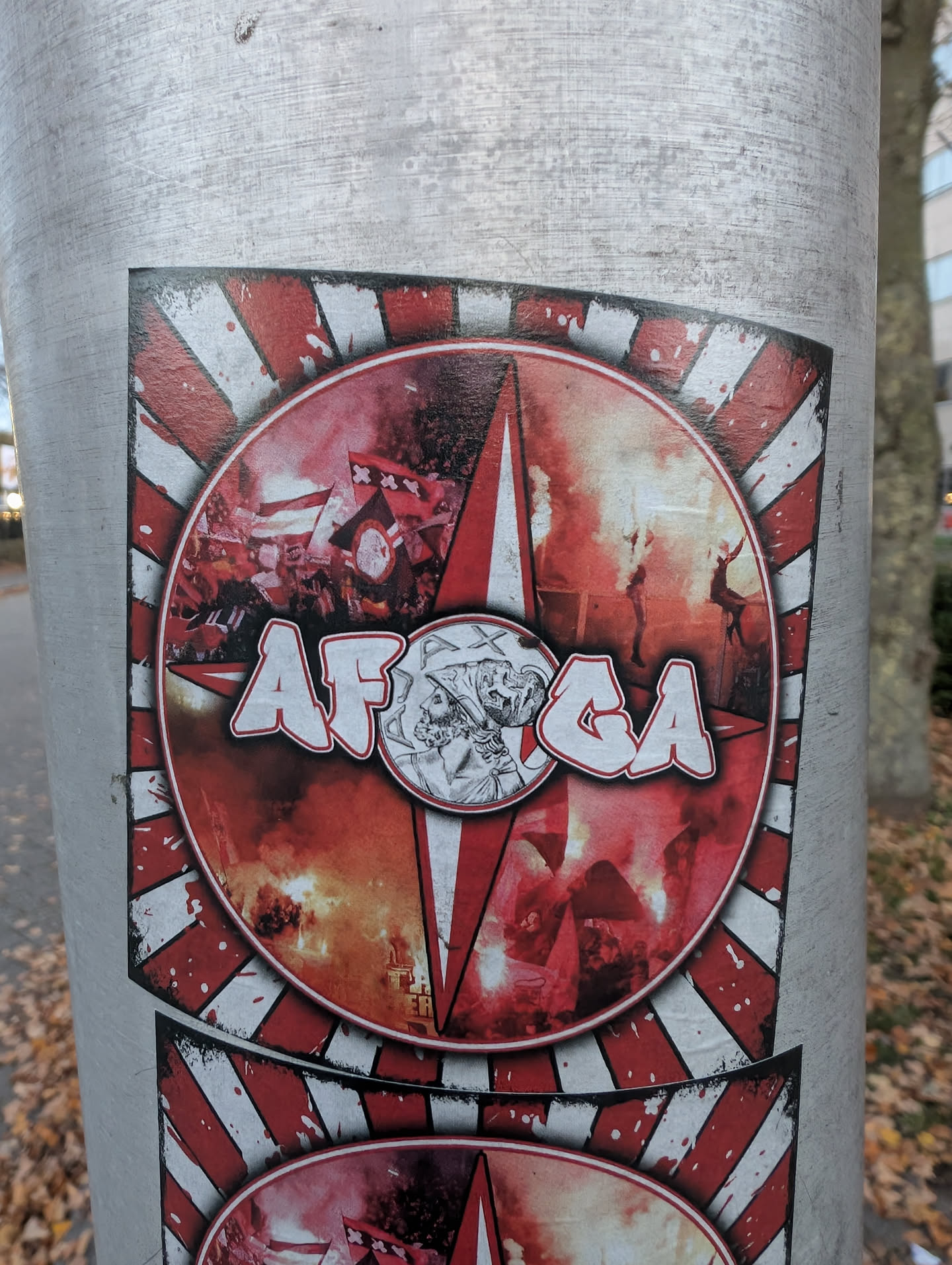 The sticker is circular with a red and white striped background radiating outwards. The center features a dynamic scene of fans waving flags and igniting flares, creating a smoky, fiery atmosphere. Overlaying this scene are the bold, white-outlined letters 'AFCA' in a graffiti-like font. To the right of the text, within a smaller circle, is a detailed monochrome illustration of a profile of a man's head, possibly a historical figure or mascot. The overall impression is one of intense fan energy and team pride.