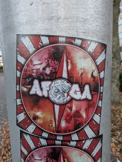 Street sticker The sticker is circular with a red and white striped background radiating outwards. The center features a dynamic scene of fans waving flags and igniting flares, creating a smoky, fiery atmosphere. Overlaying this scene are the bold, white-outlined letters 'AFCA' in a graffiti-like font. To the right of the text, within a smaller circle, is a detailed monochrome illustration of a profile of a man's head, possibly a historical figure or mascot. The overall impression is one of intense fan energy and team pride.