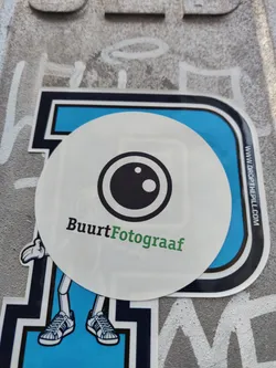 Round white sticker with a camera lens icon and the text "BuurtFotograaf" (Neighborhood Photographer) in green. It's placed on top of another sticker featuring a cartoon skeleton.