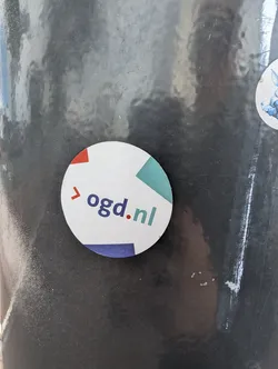 Street sticker A round sticker with the text 'ogd.nl' and a geometric design in red, teal and blue.  The sticker is affixed to a dark grey surface.